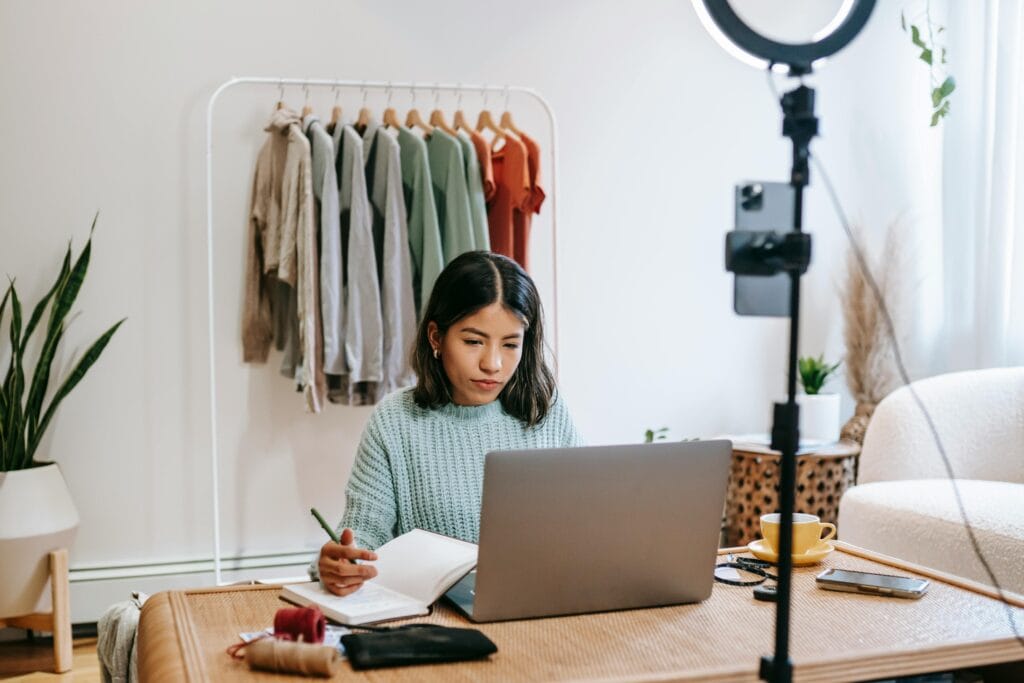pexels-photo-6347919-6347919 Focused young woman working on laptop in home office, surrounded by clothing rack and camera setup.